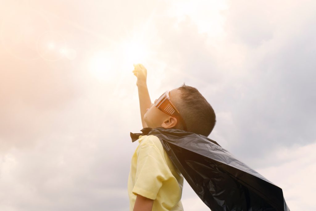 a child in a cape and sunglasses raising his fist at the sky in victory backlight by sunlight