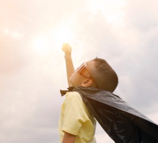 a child in a cape and sunglasses raising his fist at the sky in victory backlight by sunlight